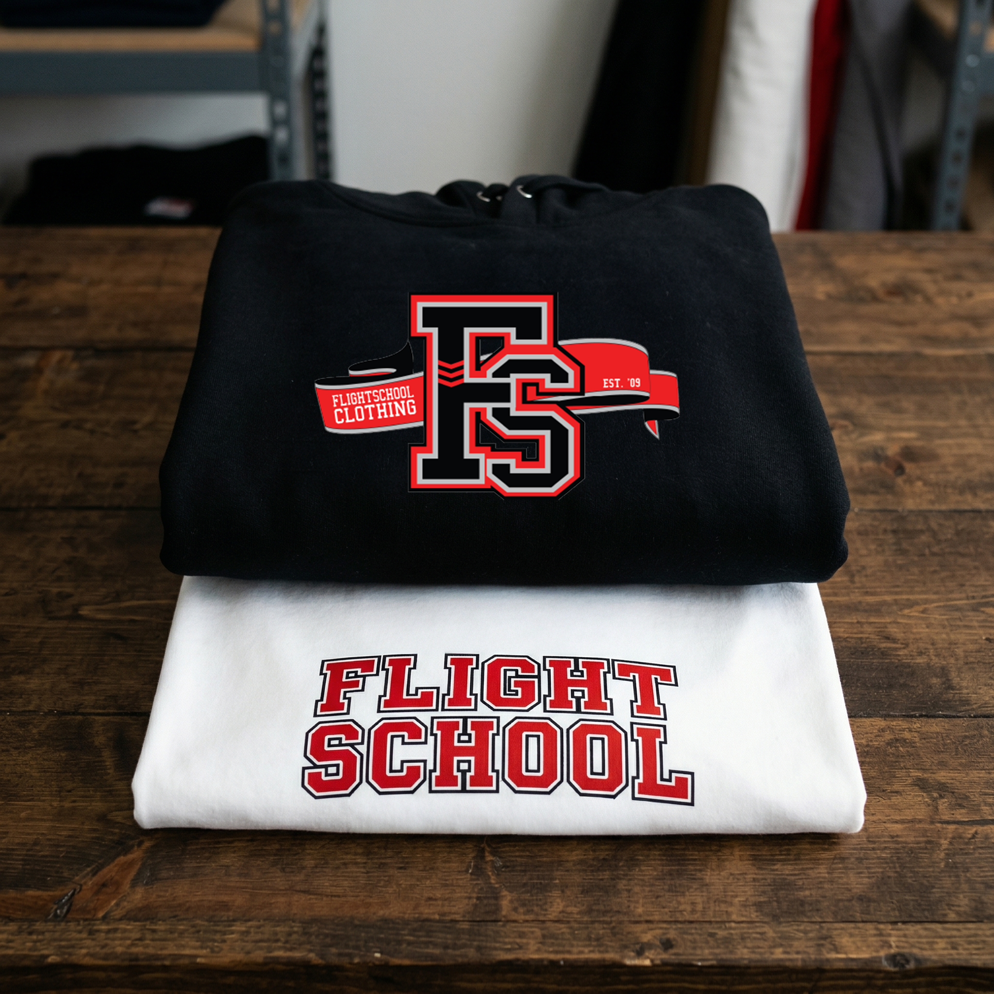 Flight School Clothing - Custom Varsity Hoodie and T-Shirt Stack A professional product stack of custom apparel for Flight School Clothing. A neatly folded black hoodie with a large red and black "FS" ribbon logo sits on top of a white t-shirt featuring "FLIGHT SCHOOL" in varsity-style lettering. The items are displayed on a dark rustic wooden table in a studio setting.